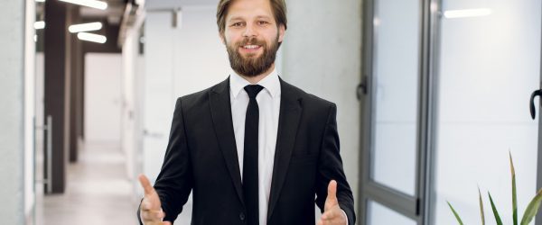 Young smiling bearded business man in elegant suit, inviting to come to the modern office. Confident